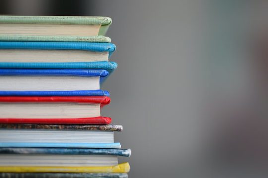 A stack of horizontal brightly-coloured green, blue, red and yellow books in the foreground, and a blurry grey background.
